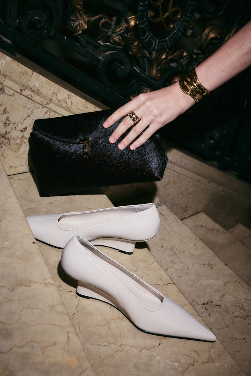 Close-up on women's fashion: off-white pointed-toe shoes, a black hair clutch in hand, and gold bracelets on stairs with black railing.