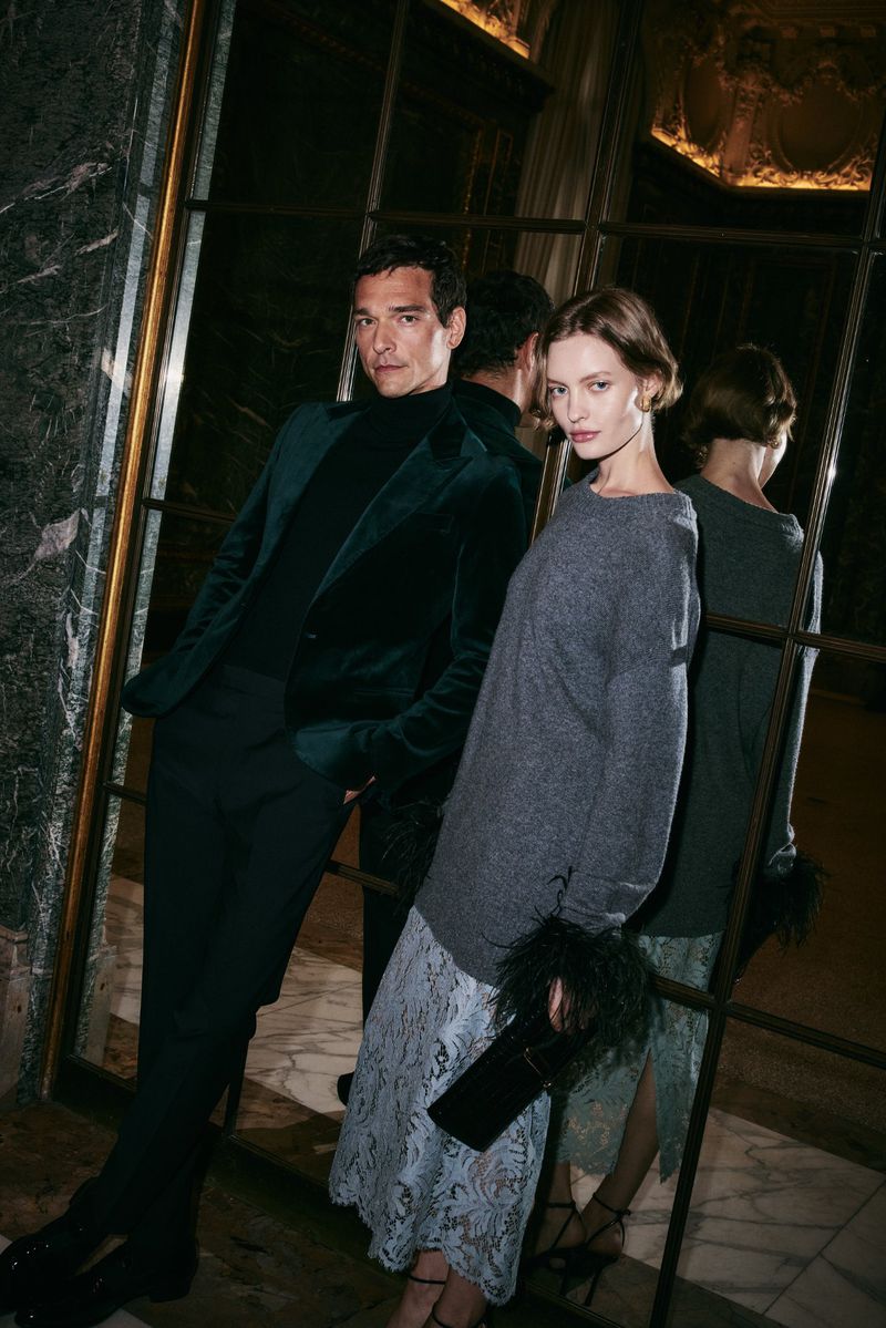 A couple poses formally. The adult male wears a green velvet suit and the adult female a gray sweater and blue lace skirt. 