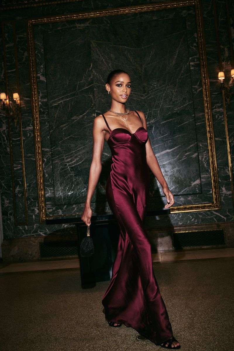 Model in a burgundy satin slip dress holding a small dark handbag inside a room with dark marble walls