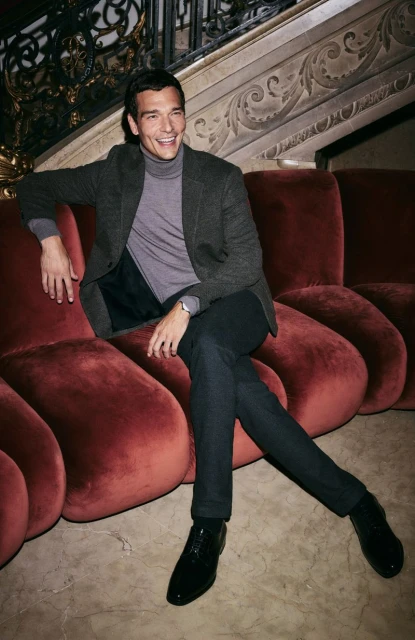 Smiling adult male wearing grey turtleneck and dark blazer sits on red modern sofa in an ornate interior setting, posing for a portrait.