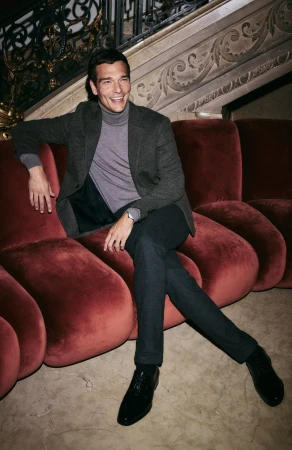 Smiling adult male wearing grey turtleneck and dark blazer sits on red modern sofa in an ornate interior setting, posing for a portrait.