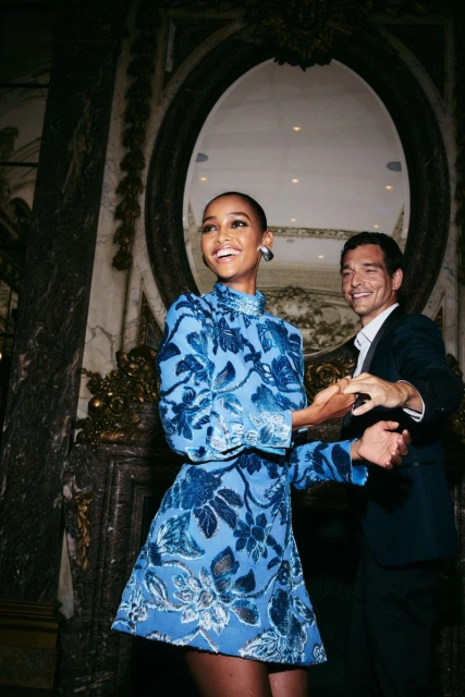 Woman in blue floral dress smiles as she holds hands with a man in a black suit, in front of an oval mirror inside a grand building.
