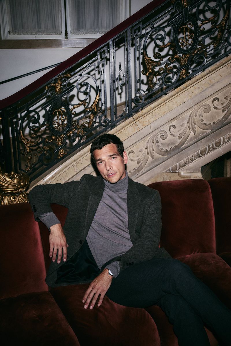 Adult man sits on a red velvet couch indoors. He is wearing a gray jacket and turtleneck with dark pants in front of an ornate staircase.