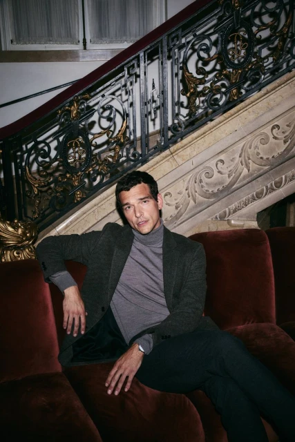Adult man sits on a red velvet couch indoors. He is wearing a gray jacket and turtleneck with dark pants in front of an ornate staircase.