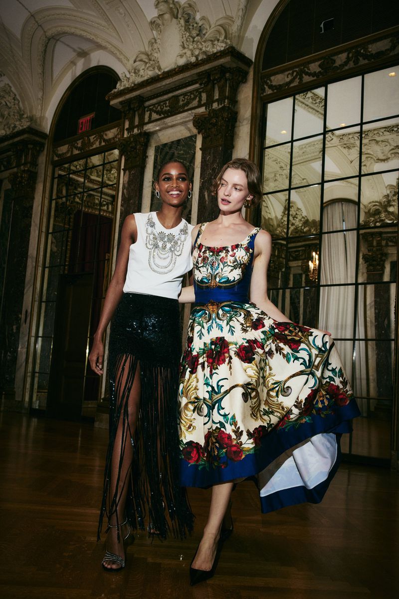 Two fashion models in a formal indoor setting, one in a white top and black fringed skirt, the other in a floral print dress.