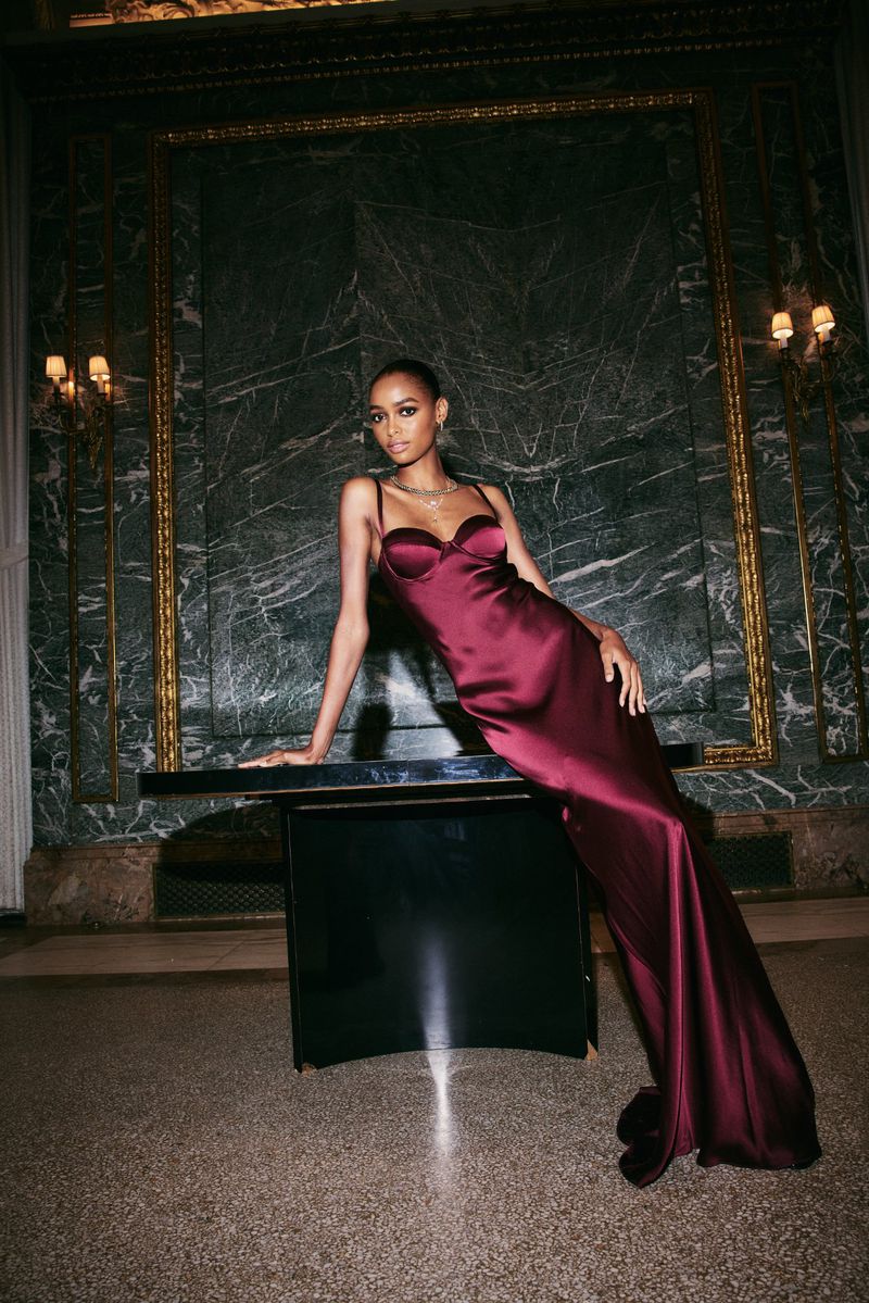 Elegant woman in burgundy satin gown poses on a black desk against a marble wall with gold trim in an indoor setting.