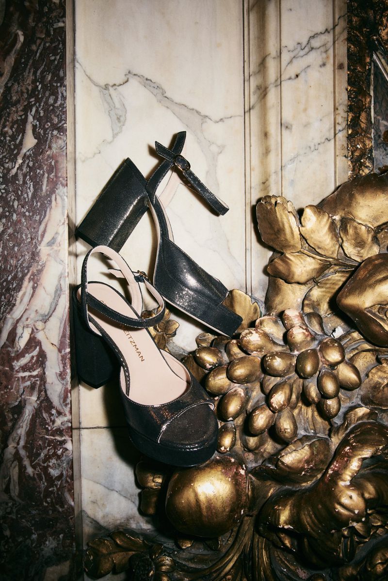 Two black high-heeled sandals are displayed against a marble and gold background, a product shot for fashion or editorial use.