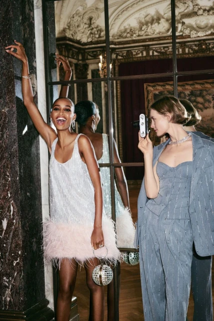 Two women pose in evening wear next to a large mirror in an ornate room, one holding a retro camera, the other raising her arm.