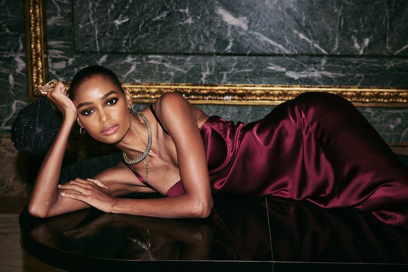 Woman in a burgundy dress poses indoors with jewelry and a beaded handbag against a marble wall with gold trim, looking at camera.