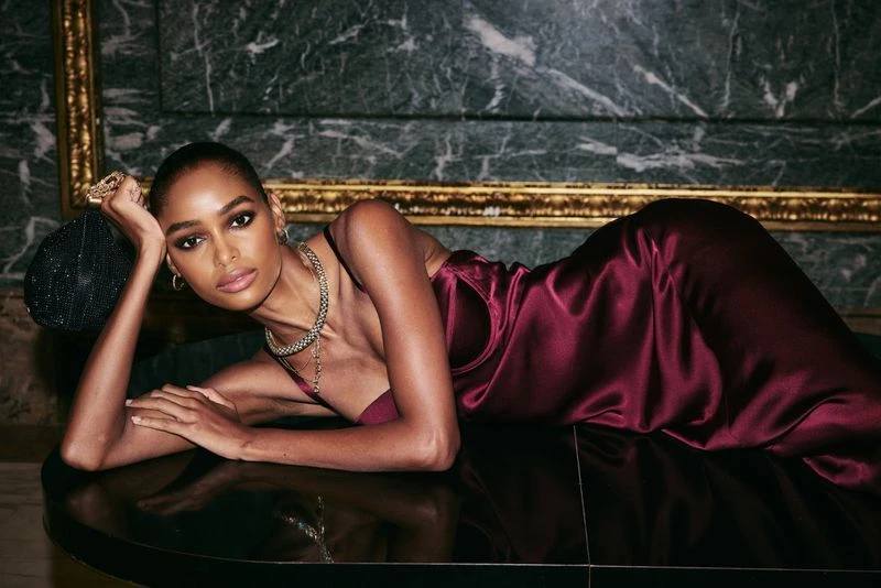 Woman in a burgundy dress poses indoors with jewelry and a beaded handbag against a marble wall with gold trim, looking at camera.