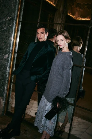 Man in dark velvet blazer and woman in grey top with feathered cuffs posing before a mirrored wall for Bloomingdale's holiday campaign