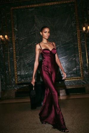 Model in a burgundy satin slip dress holding a small dark handbag inside a room with dark marble walls