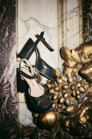 Two black platform high heels resting against an ornate gilded carving and marble wall for a holiday campaign.