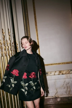 Model wearing a black coat and dress embroidered with red roses standing indoors against a classical architectural wall