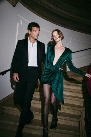 Man in a black tuxedo and a woman in a green silk dress walking down a marble staircase for Bloomingdale's holiday campaign.