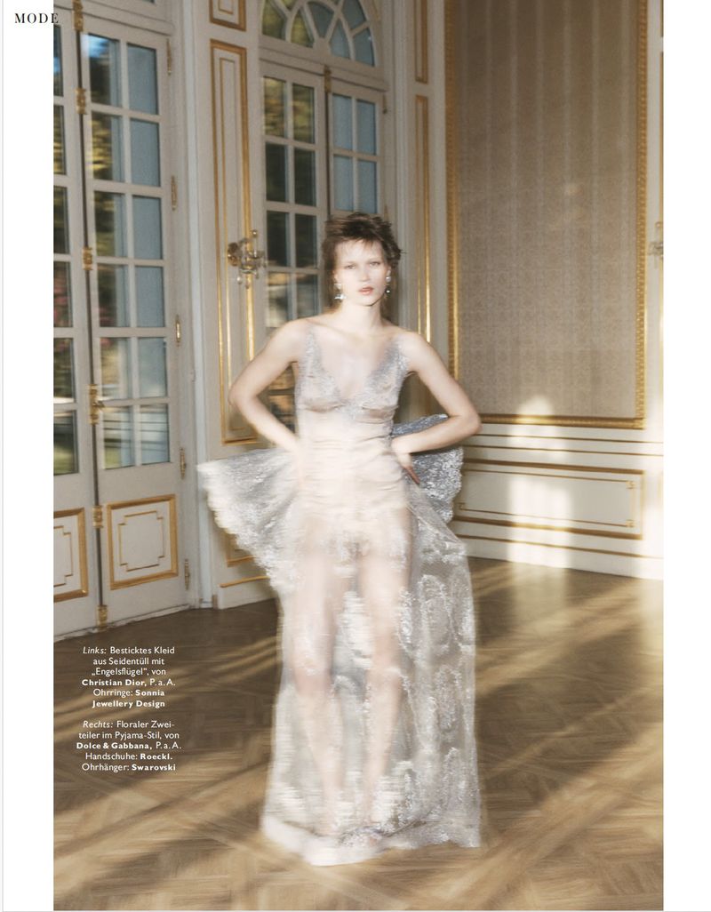 Fashion editorial shot of a woman in a sheer embellished gown posing in an ornate interior with gilded moulding and natural light.
