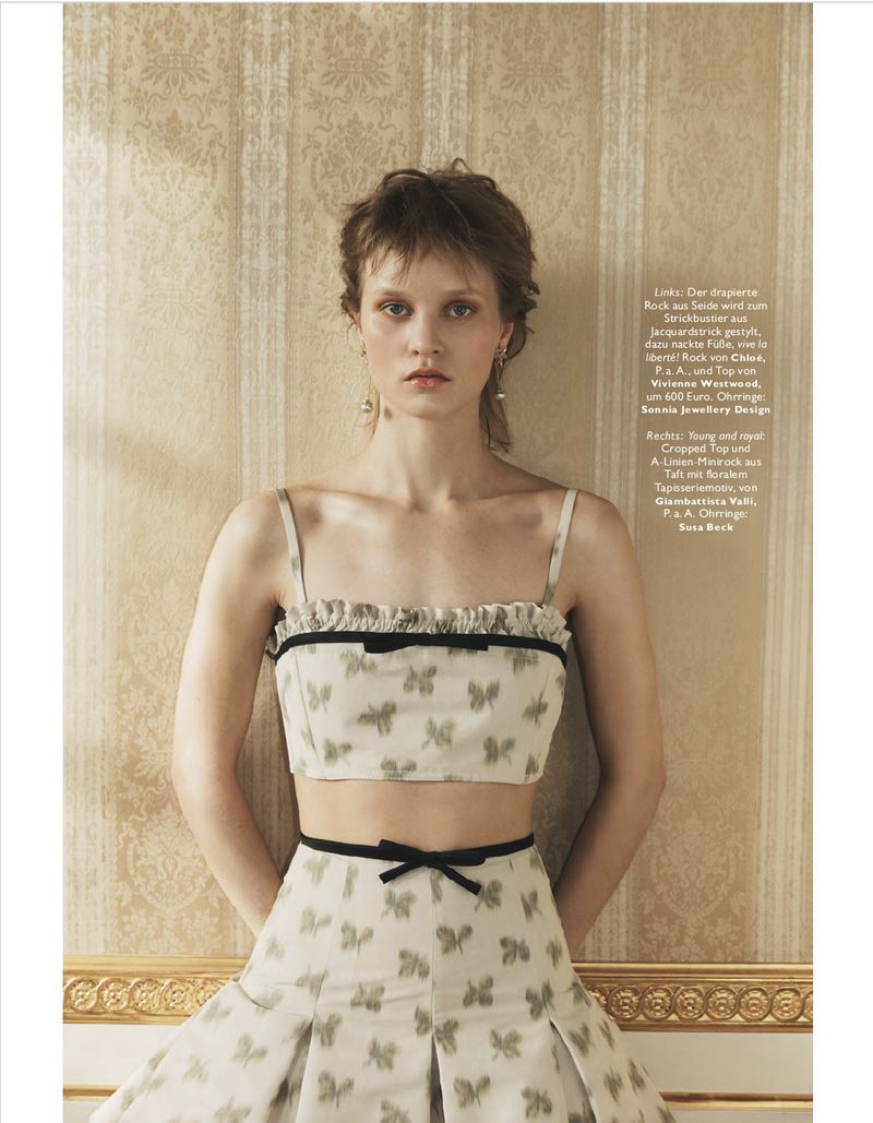 Young woman in matching crop top and pleated skirt with butterfly pattern, posing against a textured wall in a fashion editorial shot.