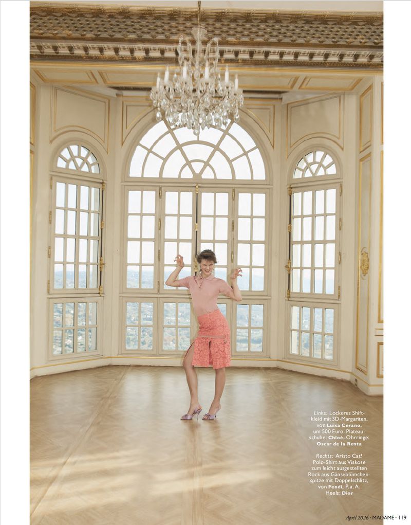A woman stands in a bright palace room, wearing a pink polo shirt and coral lace skirt, posing with her hands raised in a playful manner.