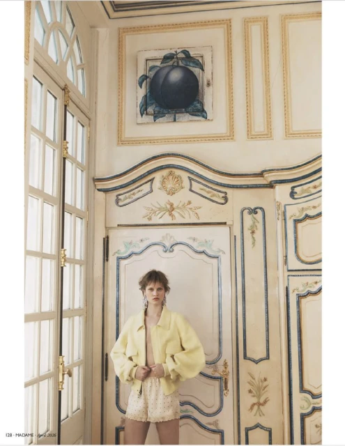 Young adult female fashion model in an indoor setting, wearing a yellow jacket and patterned shorts, posing in front of an ornate door.