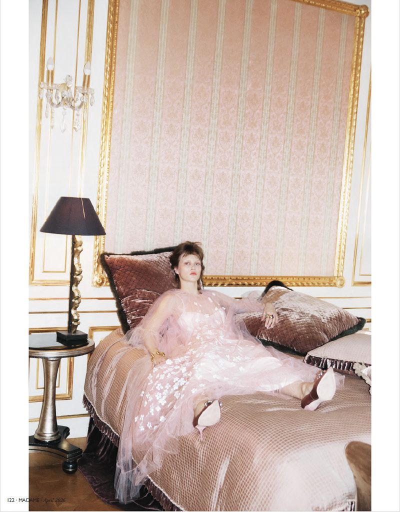 Elegant woman in a pink dress sitting on a bed in an ornate bedroom with a chandelier, decorative wall panel, and table lamp.