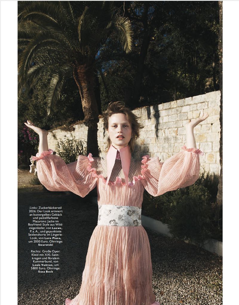 Young woman in vintage pink dress with ruffled collar and cuffs poses in front of stone wall and palm tree on sunny day.