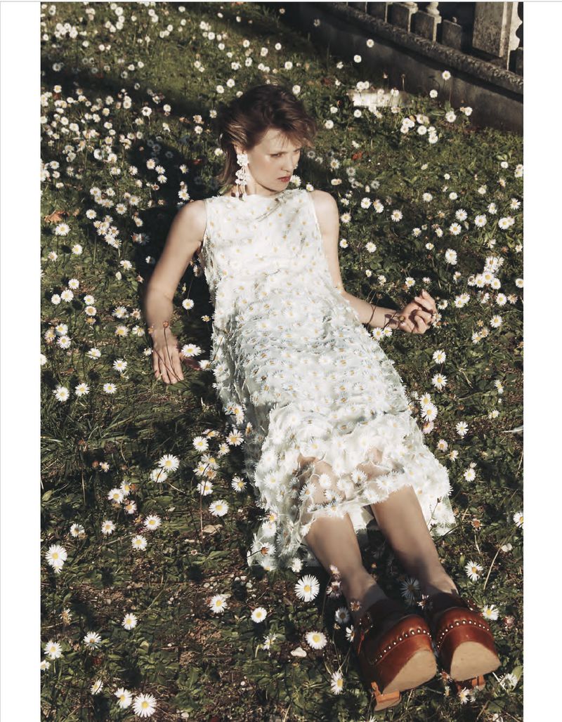 Woman in white dress lies in a field of daisies, wearing wooden platform shoes and dangling bracelet in a sunlit outdoor fashion shot.