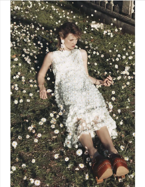 Woman in white dress lies in a field of daisies, wearing wooden platform shoes and dangling bracelet in a sunlit outdoor fashion shot.