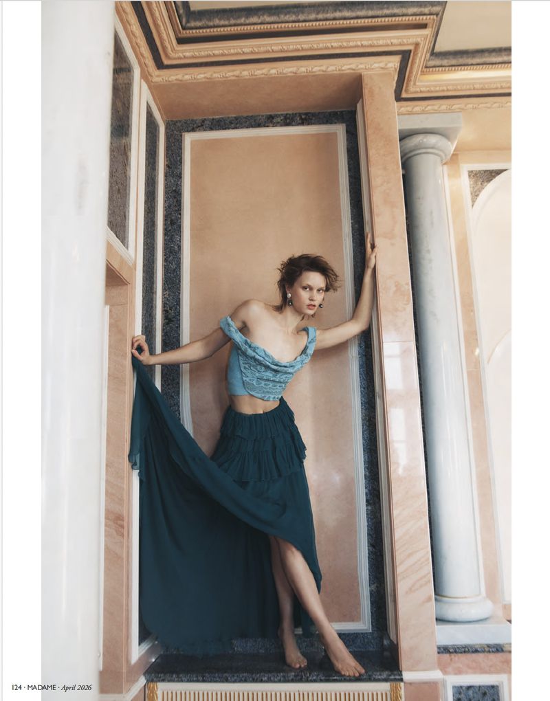 Fashion editorial shot of a barefoot young adult woman in an off-shoulder top and long skirt, leaning against a marble wall indoors.