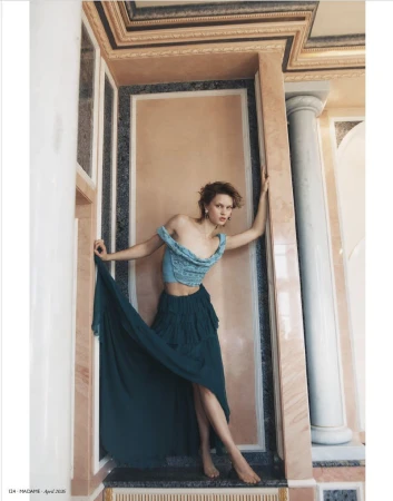 Fashion editorial shot of a barefoot young adult woman in an off-shoulder top and long skirt, leaning against a marble wall indoors.