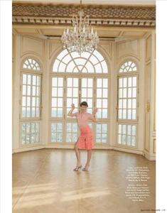 Model wearing pink top and coral skirt posing in a grand hall with arched windows and a crystal chandelier