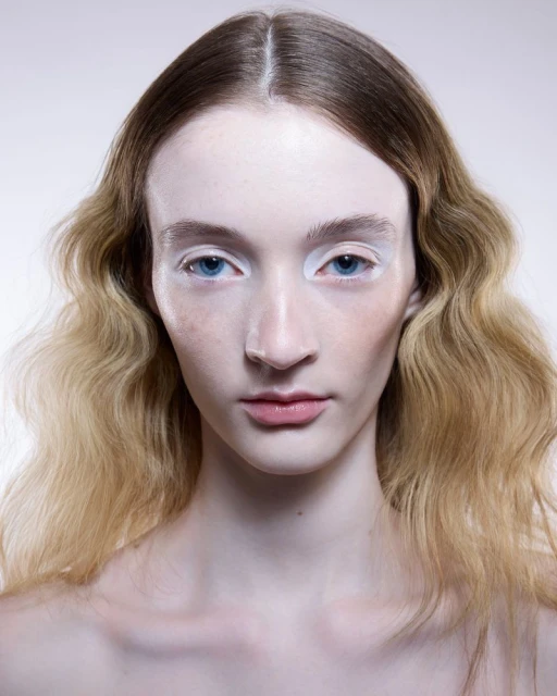 Headshot of a young adult woman with light skin and blue eyes, center parted blonde hair, light pink lipstick, pale background in studio.