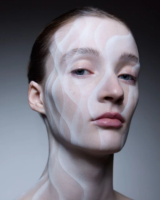Close-up portrait of a woman with white paint on her face and neck, creating wavy patterns against a dark gray background.