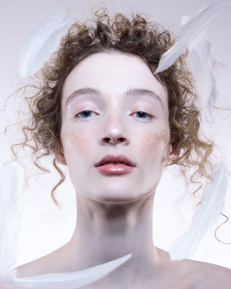 Portrait of a woman with curly hair and white feathers around her face against a pale background, capturing a soft and ethereal aesthetic.