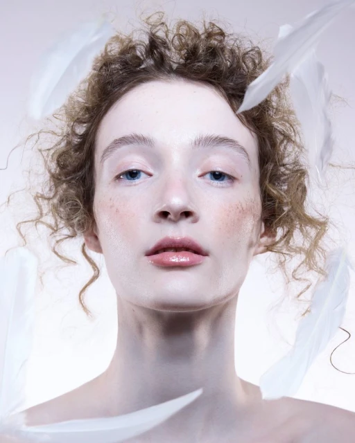 Portrait of a woman with curly hair and white feathers around her face against a pale background, capturing a soft and ethereal aesthetic.