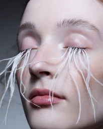Close up portrait of a woman with closed eyes wearing dramatic feather eyelashes and soft pink lipstick against a grey background.