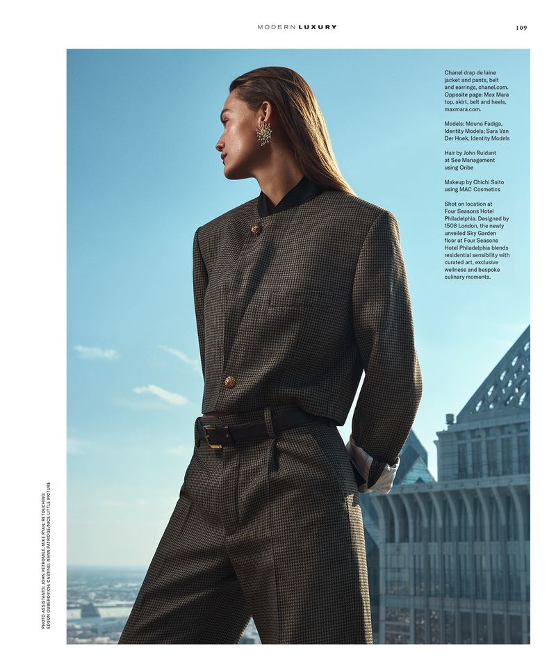 Fashion editorial shot of a woman in a suit, posing against a cityscape backdrop with a blue sky. Looking away with wet hair.