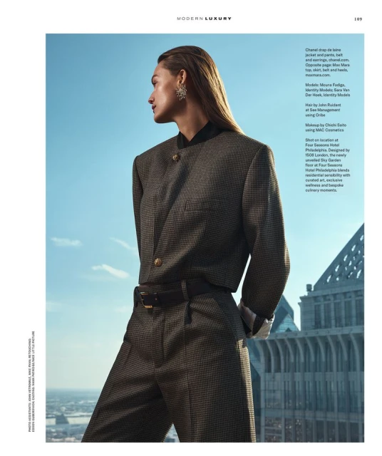 Fashion editorial shot of a woman in a suit, posing against a cityscape backdrop with a blue sky. Looking away with wet hair.