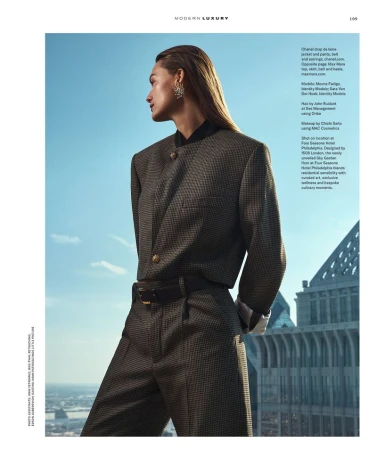 Fashion editorial shot of a woman in a suit, posing against a cityscape backdrop with a blue sky. Looking away with wet hair.