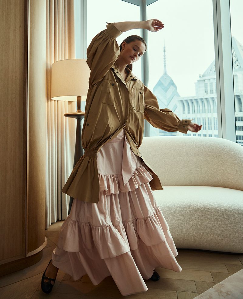Woman in tan shirt jacket and tiered pink skirt poses in front of a city view from a window, fashion editorial photography.
