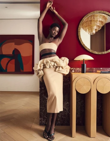 Fashionable woman poses in a beige strapless top and skirt, standing next to a console table against a red accent wall indoors.