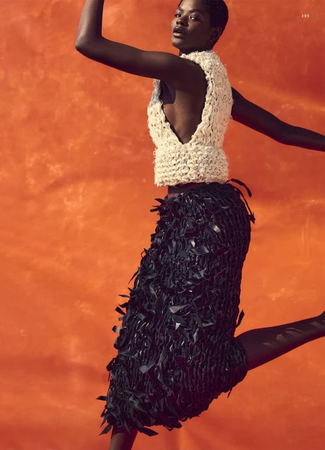 Elegant Black woman jumps with outstretched arms in sleeveless woven top and black fringe skirt against an orange mottled studio backdrop.