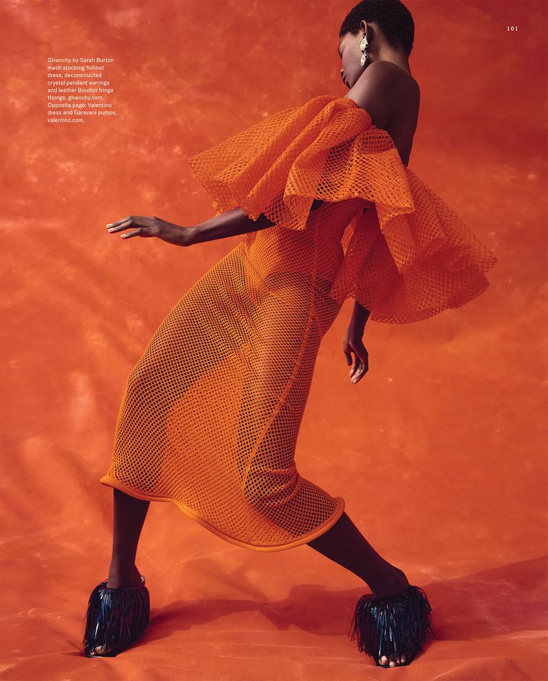 Fashion editorial shot of a Black woman in a voluminous orange mesh dress and black fringed shoes, posing against an orange backdrop.