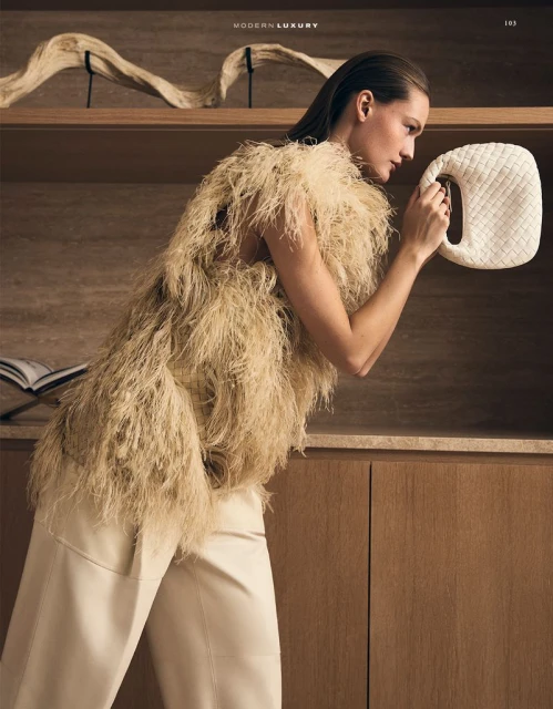 Fashion editorial shot: A woman in a feathered top and wide-leg pants holds a cream-colored woven bag indoors, against a cabinet backdrop.