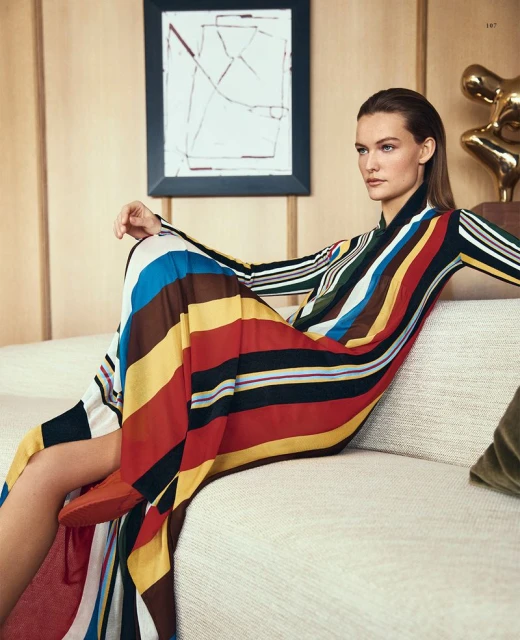 A woman poses in a long-sleeved, striped maxi dress on a beige sofa in a sunlit room with abstract artwork and modern sculpture.