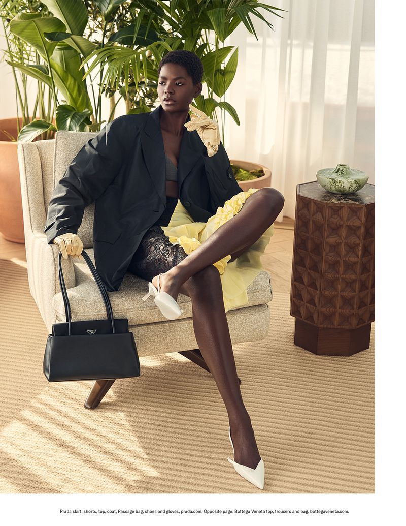 Fashion shot of Black woman in black blazer, yellow skirt, and white shoes, posing in armchair near plants, holding black purse.