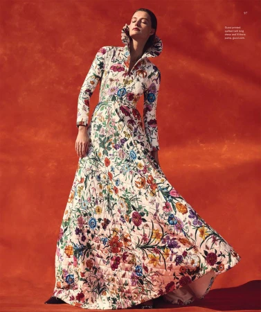 Elegant woman in floral quilted gown with ruffled collar poses against a vibrant orange backdrop in a fashion forward portrait.