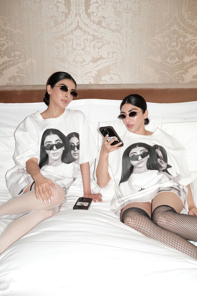 Two women pose on a bed in matching t-shirts with a black and white portrait design, fishnet stockings, and sunglasses.