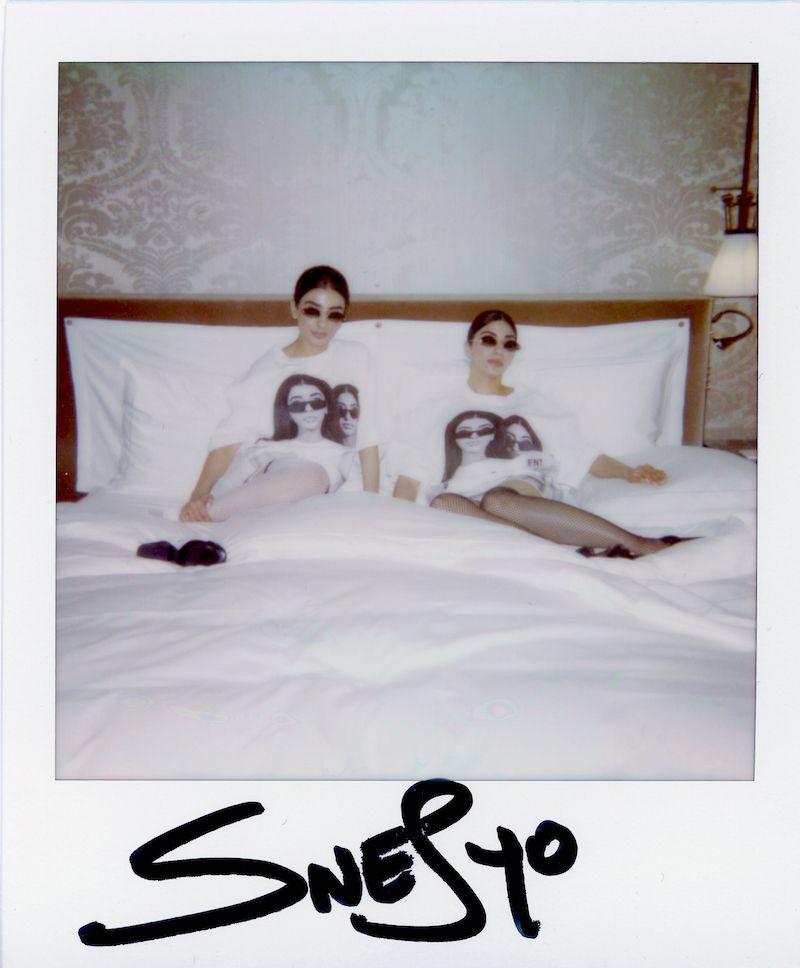 Two young women in matching graphic T-shirts lie on a white bed in a hotel room, posing for a Polaroid-style portrait with soft, diffused lighting.