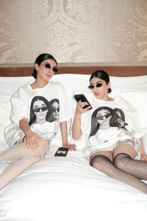 Two women wearing sunglasses and graphic t-shirts seated together on a white hotel bed for i-D Magazine