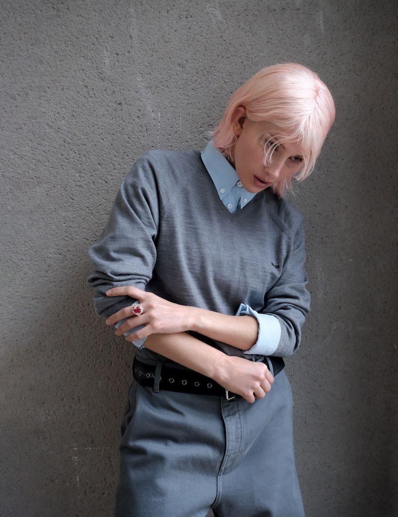 Fashionable young adult with pink hair wearing casual grey clothing, jewelry, and a black belt poses against a grey wall.